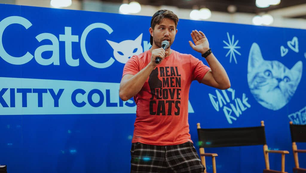 A man in a red t-shirt and backwards baseball cap speaks into a microphone in front of a blue backdrop featuring the words “CatCon Kitty Coliseum.” The man’s shirt reads “Real men love cats.”