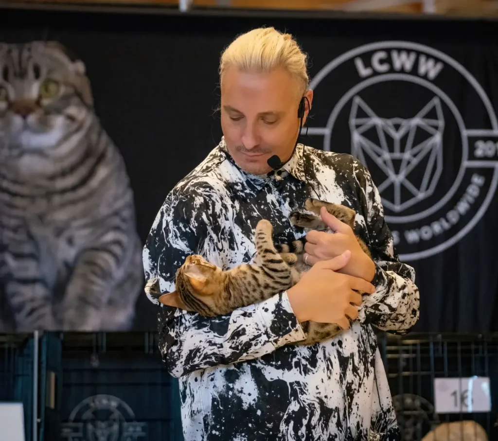 A man with a bleach blond ponytail and a black headset microphone cradles a small cat with brown-and-black stripes and spots.